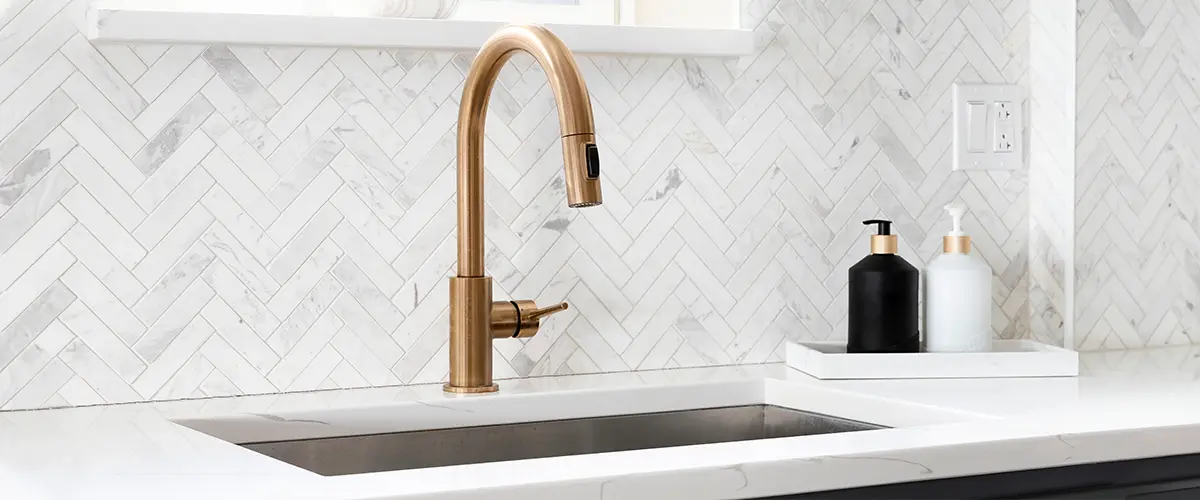 Elegant kitchen with a brass faucet, marble herringbone backsplash, white countertops, and stylish soap dispensers for a modern touch.