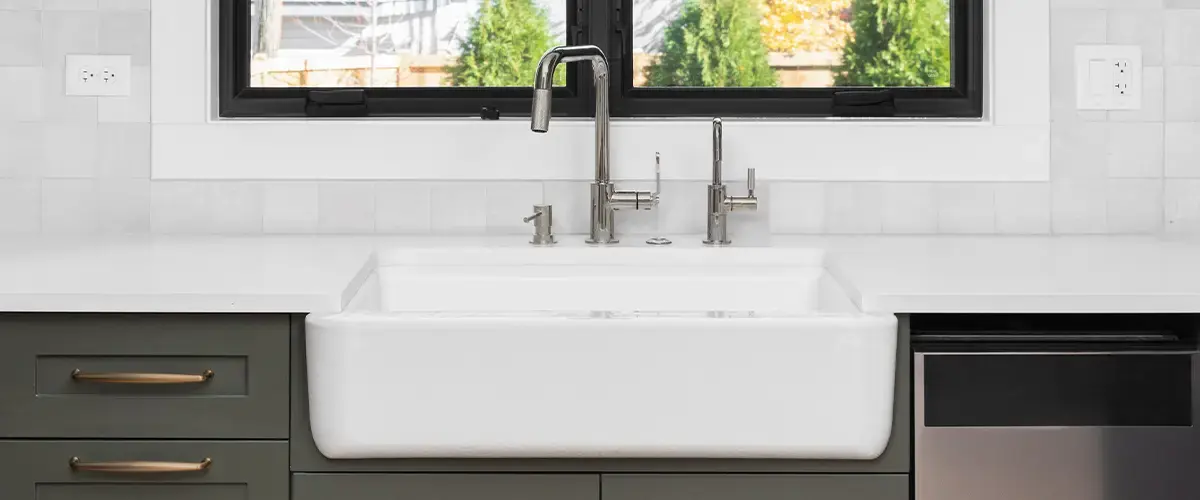 Modern kitchen featuring a white farmhouse sink, sleek stainless steel faucet, and green cabinetry with brass handles.