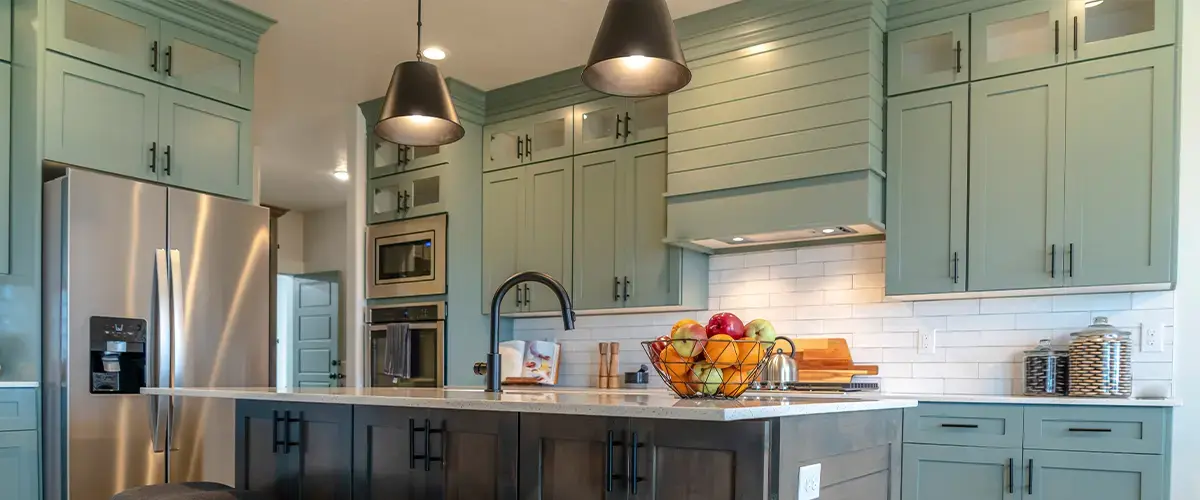 Modern kitchen featuring green cabinets, a stainless steel refrigerator, pendant lighting, and a central island with a black faucet and fruit basket.