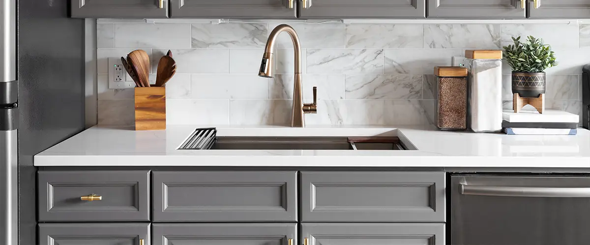 Modern kitchen with gray overlay cabinets, marble backsplash, and brass faucet over white countertop, featuring organized storage containers.