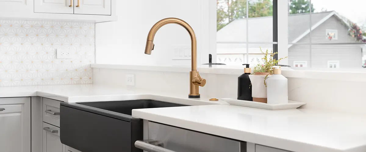 Modern kitchen with white cabinetry, a black apron sink, gold faucet, and decorative backsplash. Large window provides ample natural light. Apron Sink Vs Farmhouse Sink