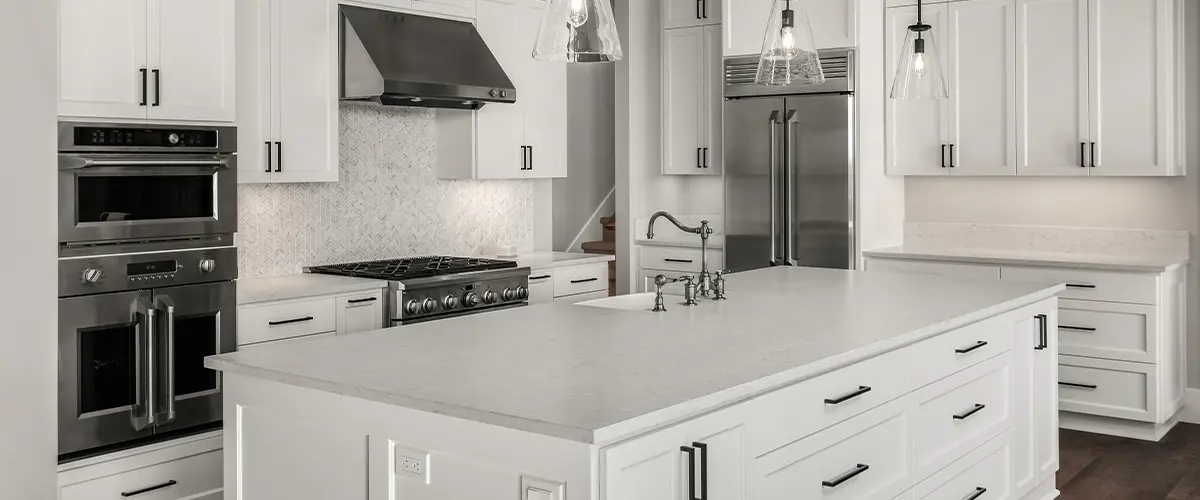 Modern white kitchen with large island, stainless steel appliances, and pendant lights, featuring sleek overlay cabinets and marble backsplash.