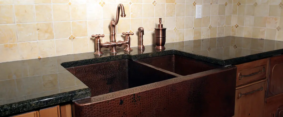 Rustic kitchen with a hammered copper farmhouse sink, matching antique copper faucet, soap dispenser, and beige tiled backsplash.