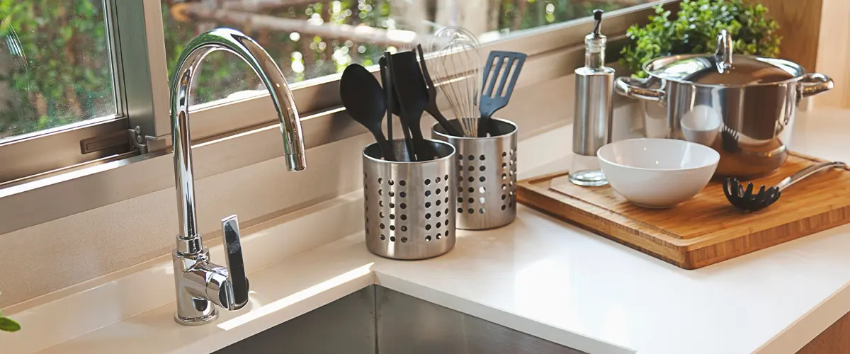 Modern kitchen sink with stainless steel faucet, cooking utensils in metal holders, and a cutting board with cookware on a white countertop