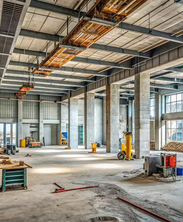 Semi-completed modern interior of a commercial building under construction, with concrete pillars, unfinished walls, and exposed ductwork, surrounded by construction equipment and tools