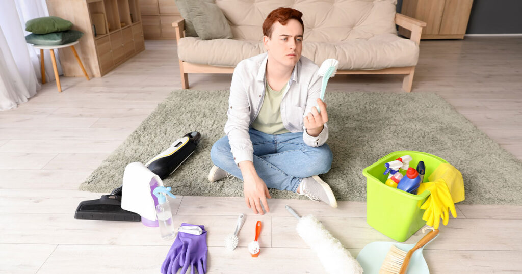 Man sitting on the floor surrounded by cleaning supplies, holding and inspecting a brush in a modern living room