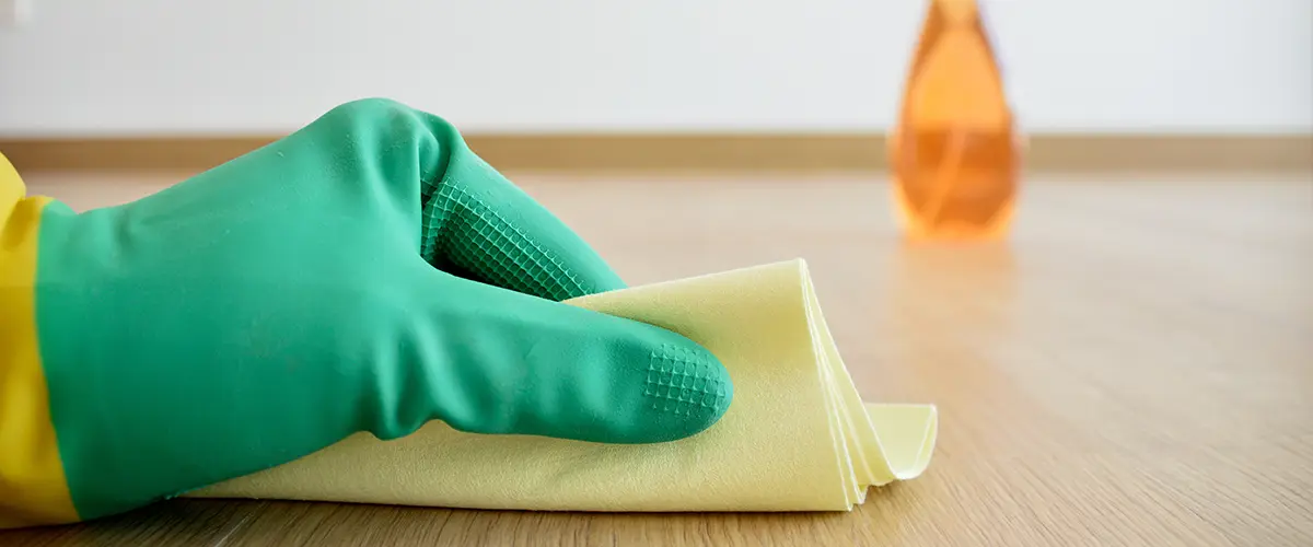 Close-up of a gloved hand cleaning a hardwood floor with a yellow cloth, with an orange spray bottle in the background