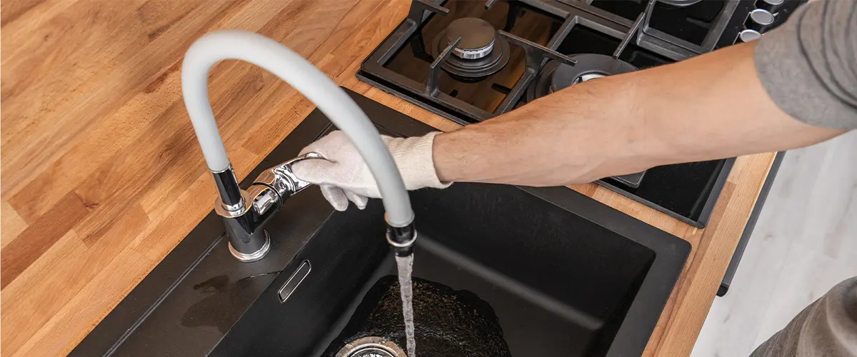 Person wearing gloves installing a faucet over a black kitchen sink with water running, wooden countertop visible.