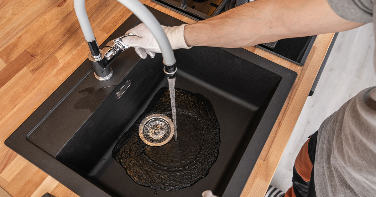 Person wearing gloves installing a faucet over a black kitchen sink with water running, wooden countertop visible.