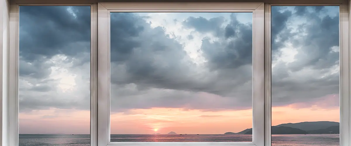 View of a serene ocean sunset through a large white-framed window, with mountains in the distance and clouds in the sky