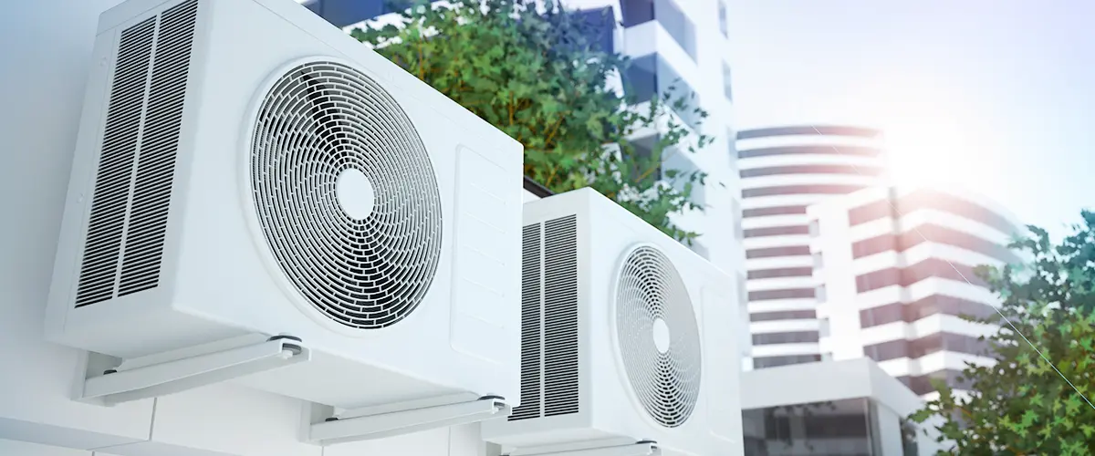 Two outdoor air conditioning units mounted on a modern building facade with city skyscrapers in the background