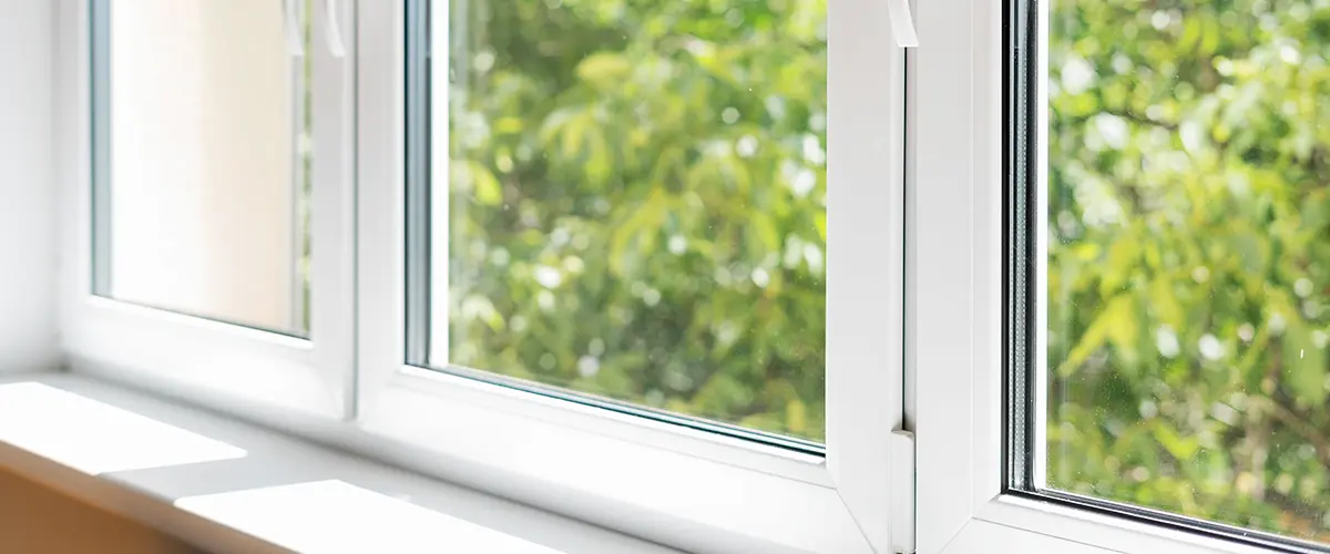 White-framed window with handles, overlooking green trees outside on a sunny day