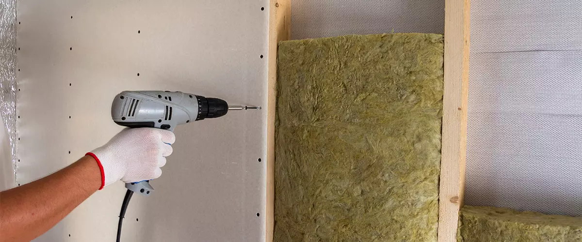 Close-up of worker hand with electric screwdriver fastening drywall to wooden timber frame with insulation mineral rock wool staff