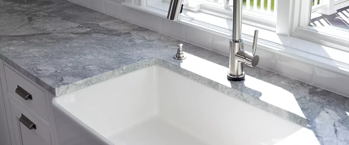 Modern kitchen sink and single handle brass faucet with soap dispenser, marble countertop and window to the garden.