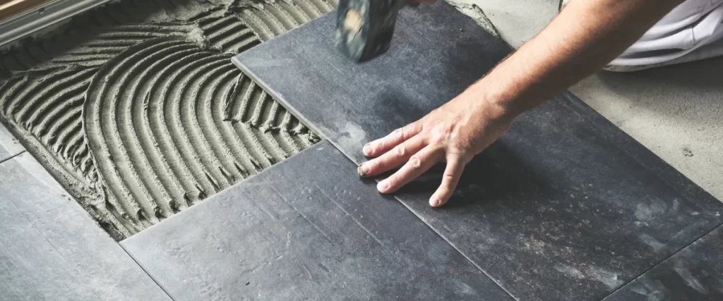 A worker installing dark-colored floor tiles using a notched trowel and mortar adhesive, ensuring proper alignment and adhesion.