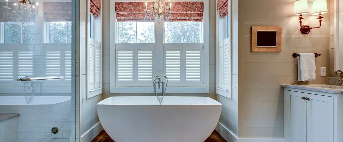 Elegant bathroom with freestanding soaking tub, chandelier, and wood flooring in front of shuttered windows.