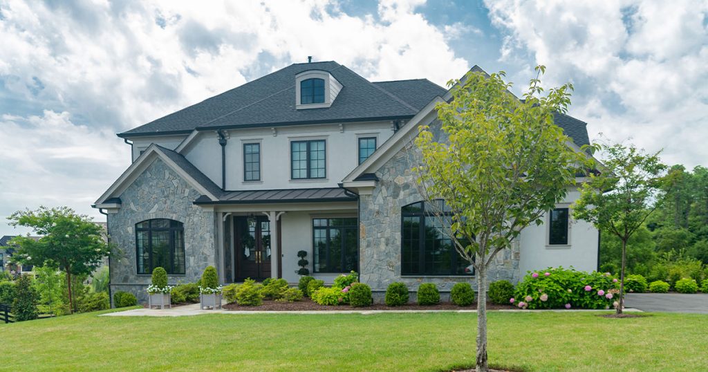 Luxury stone and stucco home with manicured lawn, modern landscaping, and dark-framed windows under a black shingle roof.