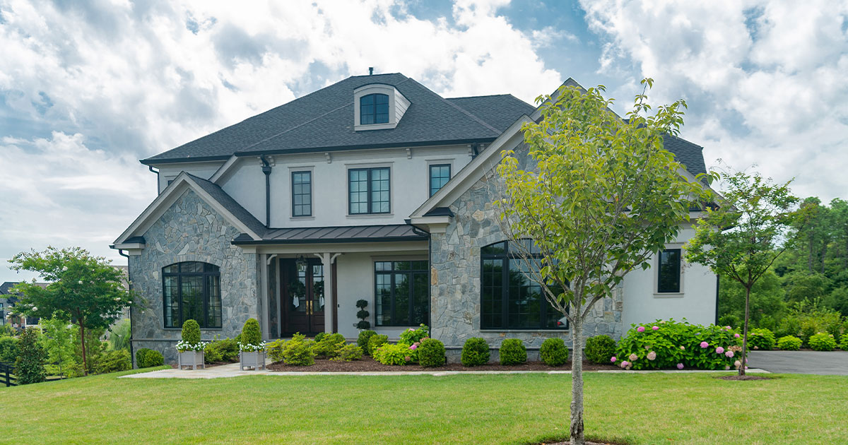 Luxury stone and stucco home with manicured lawn, modern landscaping, and dark-framed windows under a black shingle roof.